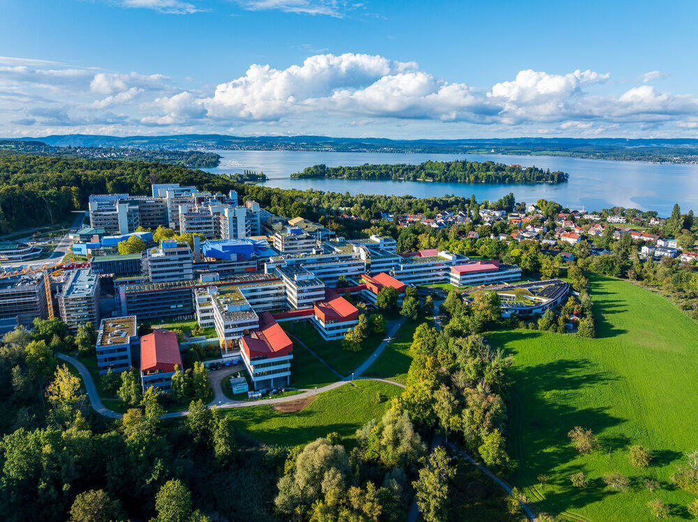 Mainau, Hockgraben Vista aérea de un campus universitario amplio y moderno, con edificios de tejados rojos, rodeado de una exuberante vegetación, junto a un lago tranquilo bajo un cielo azul.