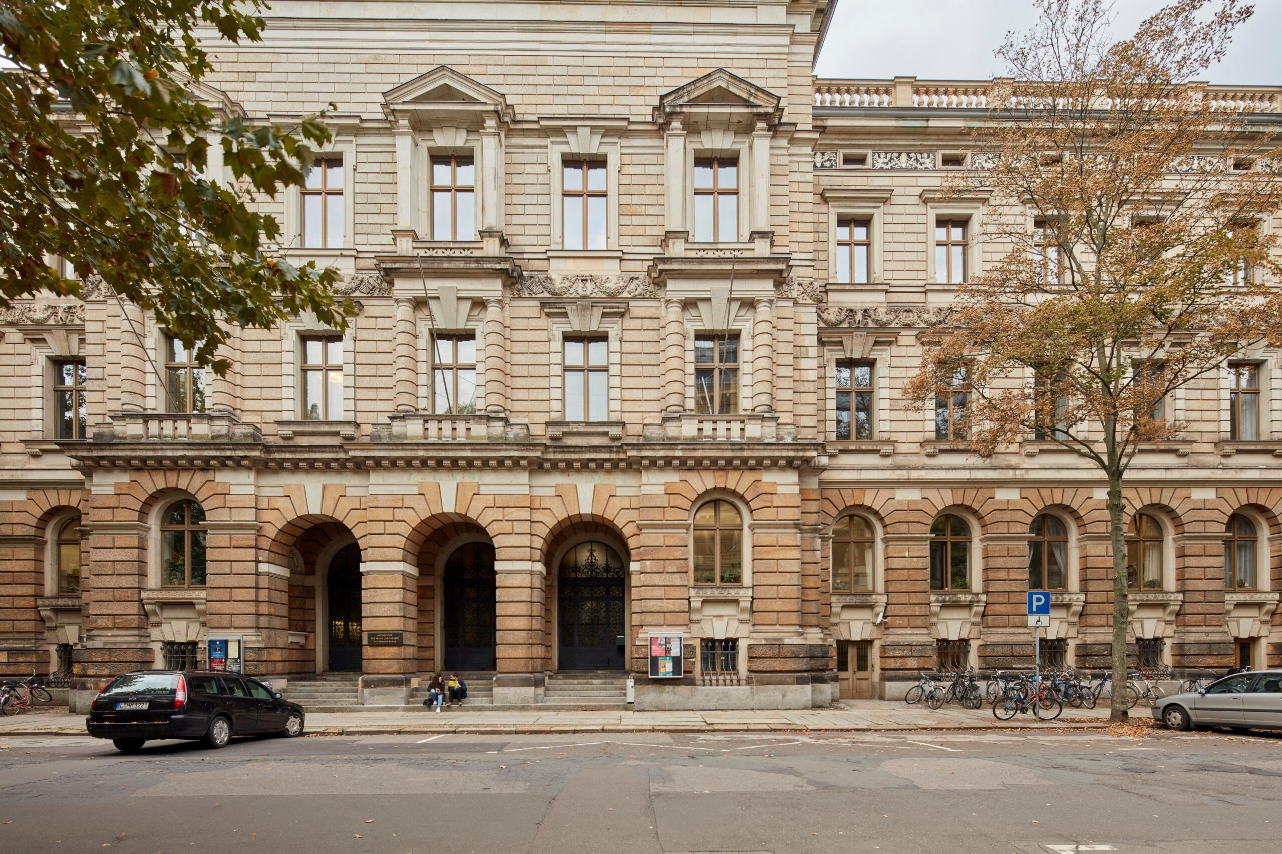 Fachada de edificio hist&oacute;rico con entrada en arco y ventanas sim&eacute;tricas. Dos coches aparcados, bicicletas alineadas y &aacute;rboles oto&ntilde;ales que a&ntilde;aden encanto al paisaje urbano.