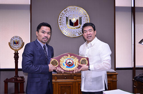 Dos hombres vestidos de gala sosteniendo un cinturón de campeón de boxeo frente al escudo del Senado de Filipinas.
